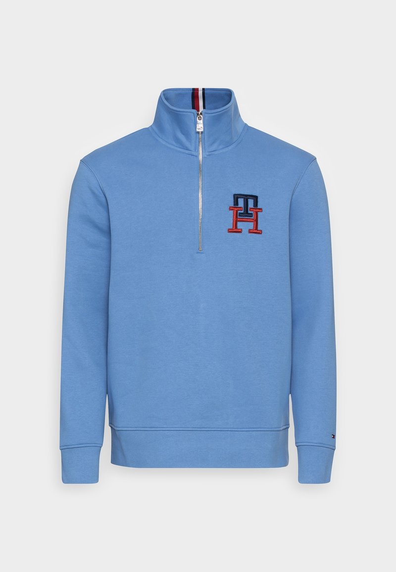 Sudadera azul claro con cremallera de un cuarto, cuello alto, logotipo "TH" en rojo y azul marino en el pecho izquierdo, y un pequeño logotipo en el puño de la manga izquierda.