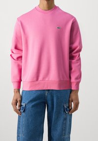 Sweat-shirt rose en tissu doux, avec un col rond, des manches longues et un petit logo vert sur la poitrine. Porté avec un pantalon cargo bleu.