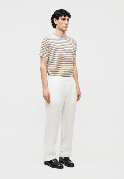 Νέος άνδρας που στέκεται, φοράει μπεζ κοντομάνικο πουκάμισο με οριζόντιες ρίγες, λευκό παντελόνι και μαύρα επίσημα παπούτσια μπροστά σε ένα απλό φόντο.