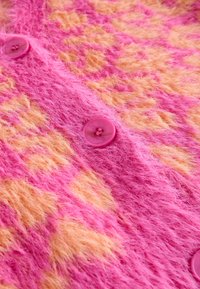 Blød lyserød og orange cardigan med et geometrisk mønster og store lyserøde knapper, der fremhæver tekstur og varme gennem sit design.