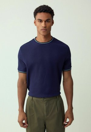 Marineblaues kurzärmeliges Strickshirt mit strukturierter Oberfläche und einem gerippten Kragen mit grünen und weißen Streifen. Kombiniert mit hellgrünen Hosen.