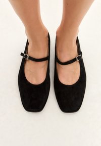 Zapatos planos de ante negro con diseño de punta cuadrada, que cuentan con una correa con una hebilla de plata sobre el empeine. Textura suave, estilo minimalista.
