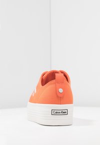 Orange canvas sneakers med vit plattformssula, bakkantens dragflik och "Calvin Klein"-logotyp på hälen. Svävande struktur, minimalistisk design.