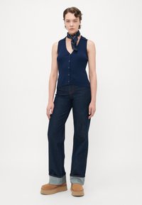 Kvinne iført en ermeløs marineblå knappet topp, vide mørke jeans med oppbrett, brune platå sko, og et marineblått skjerf knyttet rundt halsen.