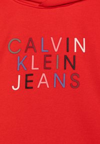 Raudona striukė su kapuci, ant kurios užrašytas "CALVIN KLEIN JEANS" tekstas įvairiomis spalvomis ir šriftais, su tekstūruotu audiniu.
