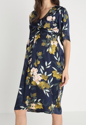 Femme enceinte portant une robe de maternité florale bleu marine longueur genou, avec des manches mi-longues et un col style portefeuille.