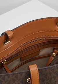 Intérieur de sac à main en cuir marron avec des accents couleur sable, comprenant plusieurs poches zippées et une étiquette de logo. Motif de logo distinctif visible.