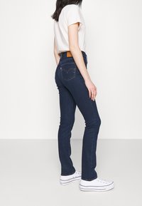 Jeans oscuros ajustados de tiro medio, con un parche de cuero en la parte posterior y costuras clásicas, combinados con zapatillas blancas.