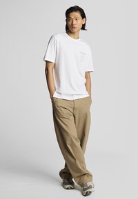 Hvid bomulds-t-shirt med korte ærmer, prydet med et lille cirkulært grafisk motiv i lyseblå. Sammen med khaki, løstsiddende bukser og grå sneakers.
