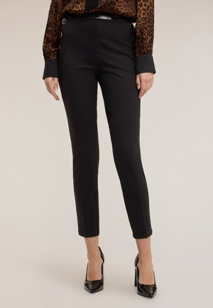 Pantalons ajustés noirs en tissu lisse, dotés de boutons sur les côtés, longueur cheville, associés à des talons hauts noirs brillants.