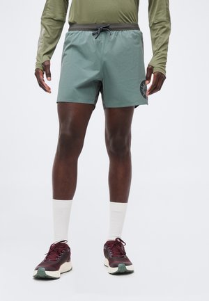 Άνδρας που φοράει πράσινα σορτσάκια με μαύρο κορδόνι, μακρυμάνικο ελιάς πουκάμισο, λευκές κάλτσες και μαρούν αθλητικά παπούτσια, στέκεται σε γκρίζο φόντο.