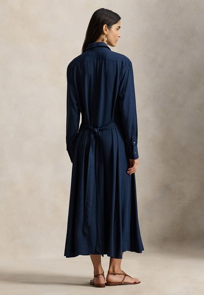 Φόρεμα πουκάμισο σε ναυτικό μπλε χρώμα με μακριά μανίκια από απαλό ύφασμα, με ζώνη στη μέση που δένει, μανσέτες με κουμπιά και φούστα που ανοίγει σε φαρδιά γραμμή. Φαίνονται επίπεδα σανδάλια.