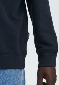 Marineblauwe sweatshirt met gestructureerde gebreide stof, geribbelde boorden en een klein label aan de zijkant. Gecombineerd met lichtblauwe spijkerbroek.