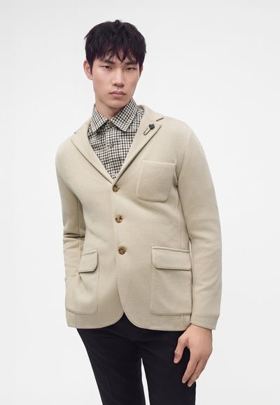 Jeune homme portant un blazer beige à trois boutons et poches plaquées sur une chemise à carreaux, posant sur un fond blanc uni.