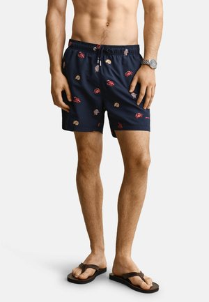 Mann trägt marineblaue Badeshorts mit roten Krabben-, Korallen- und Muschelmuster, eine Armbanduhr und schwarze Flip-Flops, steht vor weißem Hintergrund.