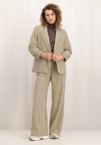 Licht olijfgroen op maat gemaakt kostuum met een enkelvoudig gebreide blazer en wijde broekspijpen. Textured donker top eronder, minimalistisch ontwerp en zachte stof.