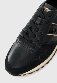 Baskets en cuir noir et en daim avec des lacets noirs, semelle blanche et noire, et logo en métal doré sur le côté et la languette.