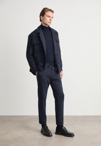 Ναυτικό μπλε υφασμάτινο μπουφάν με πολλές τσέπες πάνω από μια στενή ναυτική μπλούζα με ψηλό λαιμό, σε συνδυασμό με ταιριαστά παντελόνια και μαύρα παπούτσια. Γενικά λεία υφή.