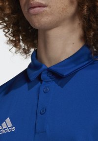 Blå polo skjorte med let stof, to knapper ved kraven og et kontrasterende sølvlogo på venstre side. Glat tekstur.