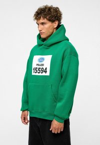 Groene hoodie met een grote buidel, voorzien van een wit rechthoekig label met "POLIZEI 15594" in zwart gedrukt.