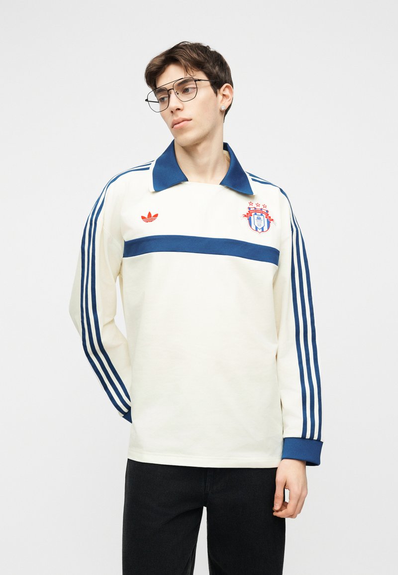 Giovane uomo con indosso una maglia sportiva a maniche lunghe bianca con strisce blu sulle maniche, colletto blu e loghi rossi, in piedi davanti a uno sfondo neutro.