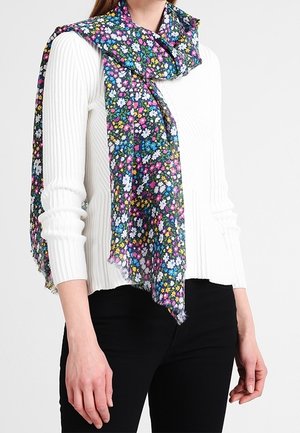 Femme portant un pull blanc à côtes avec une écharpe florale colorée drapée et nouée lâchement autour du cou, associée à un pantalon noir.
