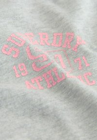 Grått tyg med en mjuk textur har ett rosa, sprucket tryck av "SUPERDRY ATHLETIC 1971" över mitten, vilket betonar bold typografi.