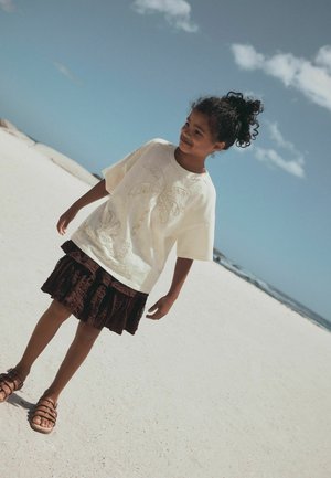 Junges Mädchen mit lockigem Haar zu einem Dutt gebunden, trägt ein weißes T-Shirt mit Palmenmotiv, einen dunklen Rock und Sandalen, steht an einem Sandstrand unter blauem Himmel.