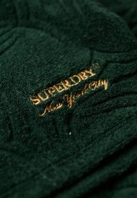 Toalla de color verde oscuro con tejido texturizado, con el logo "Superdry" bordado en dorado y detalles de un patrón de hojas. Material suave y acolchado.