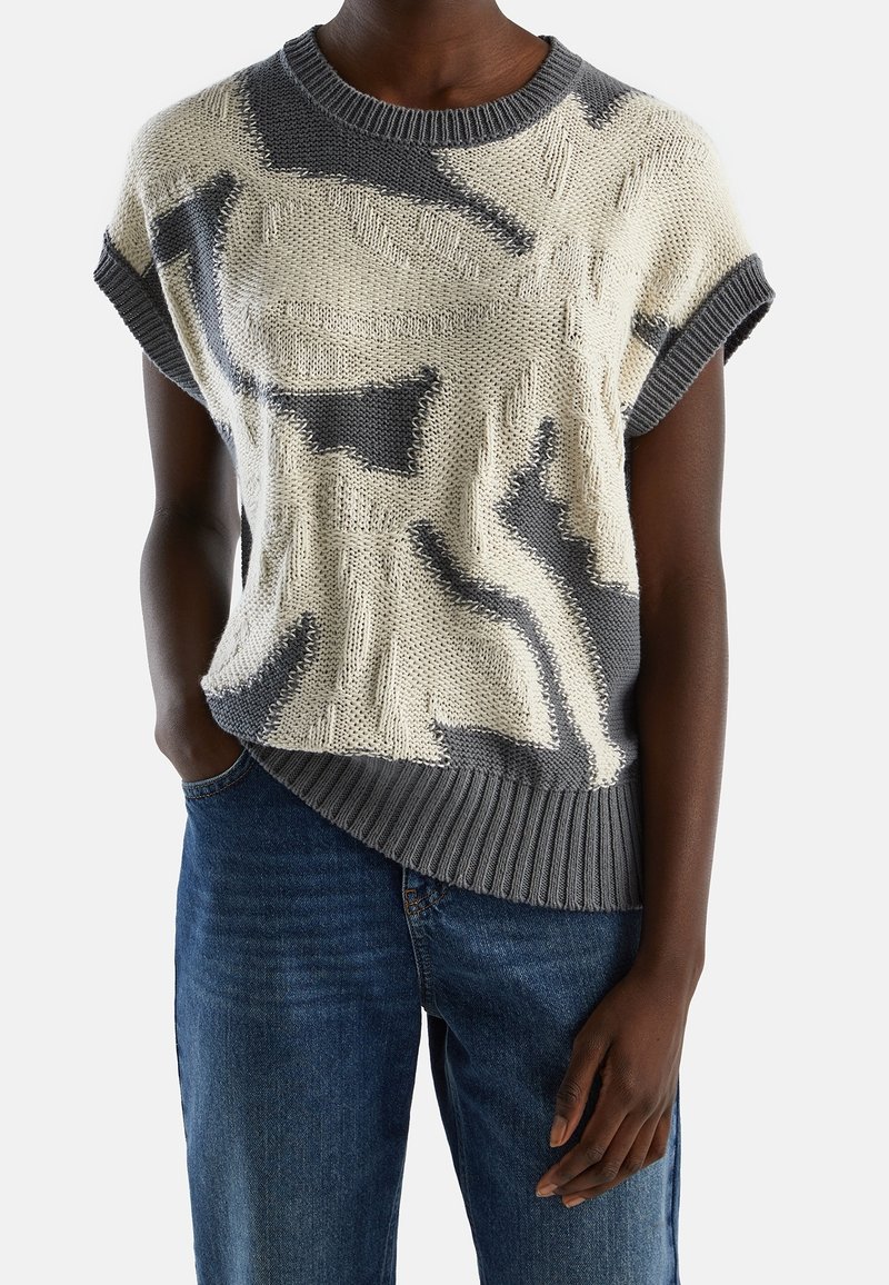 Knit sweater in cream and gray featuring a geometric pattern, short sleeves, ribbed hem, and an asymmetrical cut. Paired with blue jeans.