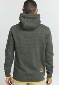 Szary hoodie z kieszenią typu kangur z przodu, z żółtymi mankietami i dołem. Haft na plecach brzmi "ISLU Since 1933". Miękka teksturowana tkanina.