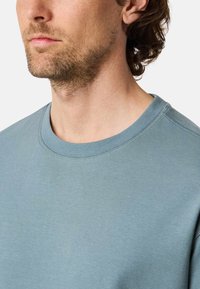 Ljusblå crewneck sweatshirt med ribbat nackparti, mjuk textur och subtila sömnadsdetaljer, synlig vid axeln och nackområdet.