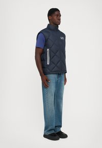 Homme portant un gilet matelassé sans manches bleu marine sur un t-shirt bleu, avec un jean et des baskets noires, se tenant devant un fond clair uni.