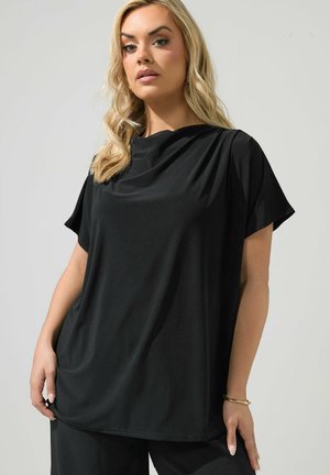 Femme blonde portant un haut noir ample à manches courtes et un pantalon assorti, posant sur un fond gris clair.