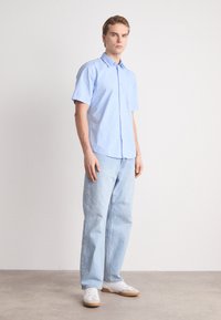 Camisa de manga corta de color azul claro con cuello abotonado, combinada con jeans de corte relajado en azul claro y zapatillas blancas. Textura de tela suave.