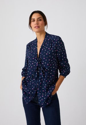 Mujer con blusa azul marino con patrón de puntos rosas y blancos, atada al frente, y vaqueros azul oscuro con las manos en los bolsillos.