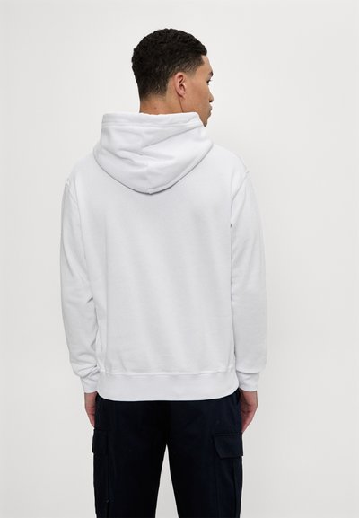 Hoodie blanc en tissu doux, avec une capuche à cordon de serrage, des poignets et un ourlet côtelés. Porté avec un pantalon cargo noir, vu de dos.
