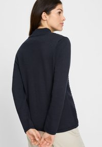 Marineblauwe cardigan met een getextureerde achterkant, geribde kraag en lange mouwen. Materiaal lijkt zacht en gebreid, met een ontspannen pasvorm.
