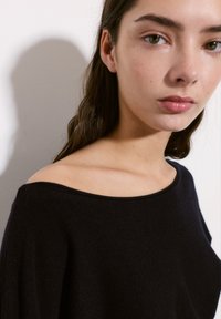 Črn, mehak pulover s širokim izrezom in dizajnom brez naramnic. Tkanina ima rahlo teksturo; rokavi so nekoliko ohlapni.