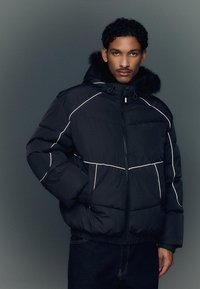 Musta untuvatakki valkoisilla saumoilla, jossa on huppu ja tekoturkisreunus, vetoketjusuljenta ja joustavat hihansuut. Lämmin, tikattu rakenne.