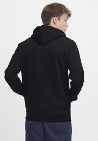 Sudadera negra con una textura suave y bolsillo tipo canguro. Cuenta con capucha con cordón y puños acanalados. La vista trasera muestra un diseño sin costuras.