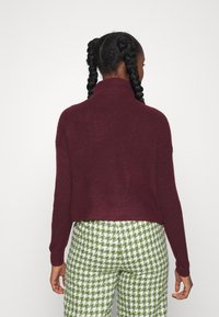 Una mujer con trenzas lleva un suéter de canalé color granate y pantalones con un estampado de pata de gallo en verde y blanco, de espaldas a la cámara.