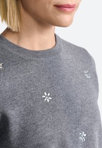 Grijze trui van zacht materiaal, met decoratieve bloembroderie en heldere rhinestones op de schouders en borstpartij.