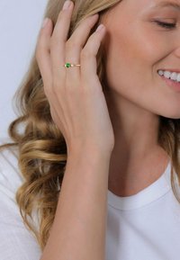 Bague en or avec une pierre précieuse verte en forme de cœur, flanquée de petites pierres ; portée sur une main avec des cheveux bouclés, vêtue d'un haut blanc.