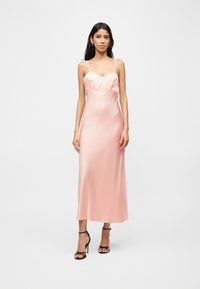 Naine, kes kannab varrukateta heleroosat satiinist kleiti õhukeste rihmadega ja musti kõrgete kontsadega sandaale, seisab ühevärvilise tausta ees.