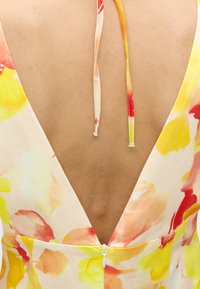 Vue de dos d'une personne portant une robe sans manches avec un dos en V profond et un motif floral dans des tons jaunes, rouges et orangés.