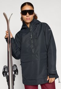 Črna vodoodporna smučarska jakna s sprednjim žepom, polovičnim zadrgo in kapuco. Model drži smuči z črnimi vezmi proti nevtralni ozadju.