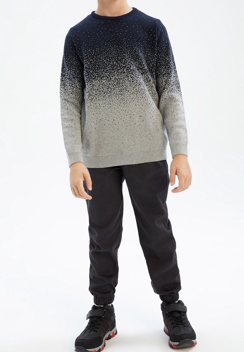 Enfant portant un pull à dégradé du noir au gris tacheté, un pantalon de jogging noir et des baskets noires aux semelles rouges, debout sur un fond blanc.