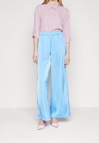 Woman wearing loose light blue satin pants, a lavender button-up shirt with pocket, and light pink high heels, standing against a white wall.