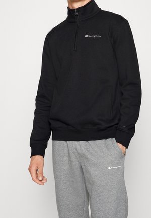 Mann trägt schwarzen Sweatshirt mit Viertel-Reißverschluss und graue Jogginghose, beide mit weißen Champion-Logos auf der Brust und dem Oberschenkel.